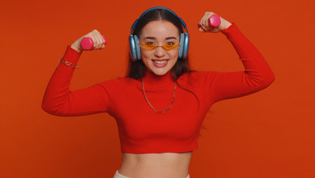 Young fit woman listening music via headphones, working out, pumping up arm muscles lifting pink...