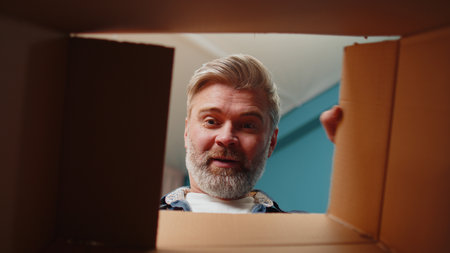Happy surprised winner man unpacking delivery parcel at home. Smiling satisfied senior guy shopper online shop customer opening cardboard box receiving purchase gift headphones by fast postal shippingの写真素材