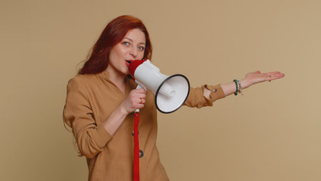 Redhead woman talking with megaphone, proclaiming news, loudly announcing advertisement, warning using loudspeaker to shout speech, pointing empty place. Copy-space. Young girl on beige backgroundの写真素材