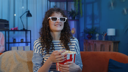 Excited young woman sits on sofa eating popcorn snacks and watching interesting TV serial, sport...