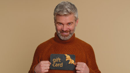 Senior mature man showing pointing on gift discount certificate voucher coupon for store holidays sale. Middle-aged guy with shopping christmas surprise card isolated alone on beige studio backgroundの写真素材