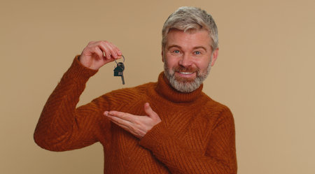 Middle-aged old man real estate agent lifting hand showing the keys of new home house apartment, buying or renting property, mortgage loan. Senior mature guy isolated alone on beige studio backgroundの写真素材
