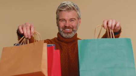 Middle-aged old man showing shopping bags, advertising discounts, smiling looking amazed with low...