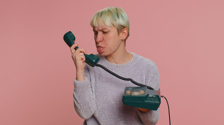 Crazy millennial woman with short hair talking on wired vintage telephone of 80s, fooling, making silly faces. Young girl isolated alone on pink studio wall background. Lgbt gay lesbian people conceptの写真素材