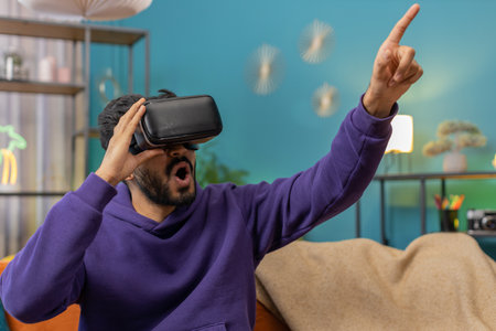 Indian man using virtual reality futuristic technology VR app headset helmet to play simulation 3D 360 video game, watching film movie at modern home apartment. Hindu guy in goggles sitting on couchの写真素材