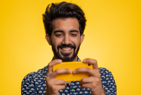 Worried funny addicted indian man enthusiastically playing racing video games on mobile phone. Handsome young guy using smartphone gadget app with drive simulator isolated on yellow studio backgroundの写真素材