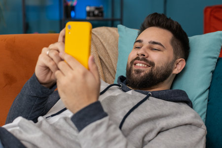 Happy man lying on couch uses mobile phone smiles at home living room apartment. Caucasian guy texting share messages content on smartphone social media applications online, watching relax movieの写真素材