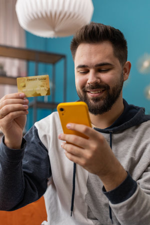 Caucasian man using credit bank card and smartphone while transferring money, purchases online shopping cashless order food delivery at home apartment indoors. Happy young guy sitting in room on couchの写真素材