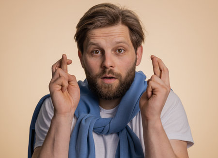 Young man crossed fingers looking at camera asking for good luck news, wishing good education exam results, dreaming about win victory in lottery jackpot. Guy isolated on beige studio backgroundの写真素材