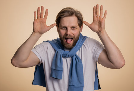 Comical funny young caucasian man making playful silly facial expressions and grimacing, fooling around, showing tongue. Handsome crazy adult guy bullying isolated on studio beige background indoorsの写真素材