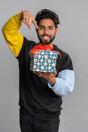 Positive smiling young indian man presenting birthday gift box stretches out hands, offer wrapped...