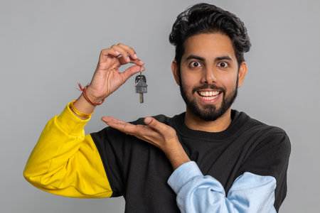 Young indian man real estate agent lifting hand showing the keys of new home house apartment, buying or renting property, mortgage loan. Bearded hindu guy isolated on gray studio background, indoorsの写真素材