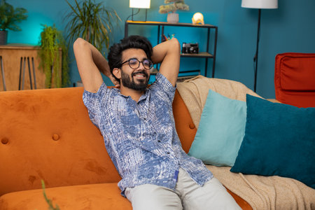 Tired indian man lying down on comfortable couch, taking a rest, daytime nap. Portrait of carefree hindu guy holding hands behind head relaxing, falling asleep at home apartment living room on sofaの写真素材