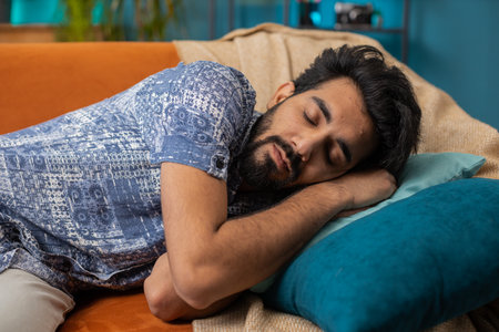 Tired indian man lying down in bed taking a rest. Carefree young hindu guy napping, falling asleep on comfortable sofa with pillows at home apartment room. Closed his eyes enjoy daytime nap aloneの写真素材