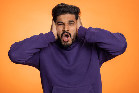 Dont want to hear and listen. Frustrated annoyed irritated indian man covering ears gesturing No, avoiding advice ignoring unpleasant noise loud voices. Handsome hindu guy alone on orange backgroundの写真素材