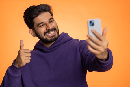 Indian man traveler blogger, taking selfie on smartphone, communicating video call online with family or friends, live translation stream. Handsome hindu guy isolated on orange background, indoorsの写真素材