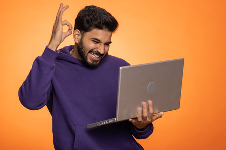 Excited happ young indian man typing on laptop computer, working on project, enjoying results, winning lottery, celebrating success online shopping, win, okay. Hindu guy isolated on orange backgroundの写真素材