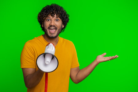 Indian man talking with megaphone, proclaiming news, loudly announcing advertisement, warning using loudspeaker to shout speech, pointing empty place. Copy-space. Handsome guy on chroma key backgroundの写真素材