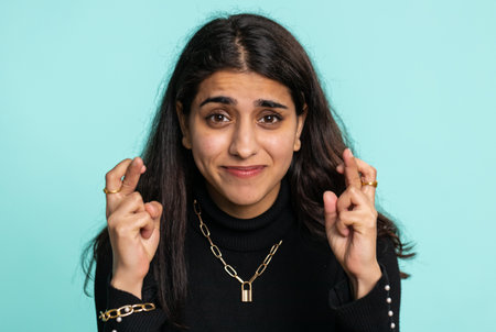 Indian young woman crossed fingers looking at camera asking for good lucky news, wishing good exam results dreaming about win victory in lottery jackpot. Hindu girl isolated on blue studio backgroundの写真素材