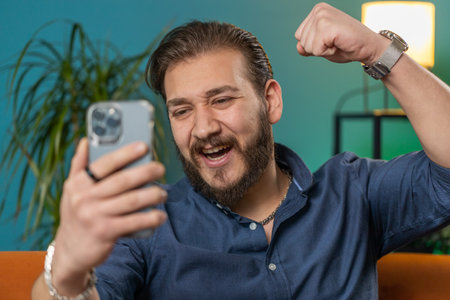 Happy lebanese man use mobile smartphone typing browsing say Wow yes found out great big win good news celebrate lottery jackpot doing winner gesture. Arabian young guy at home in room sitting on sofaの写真素材