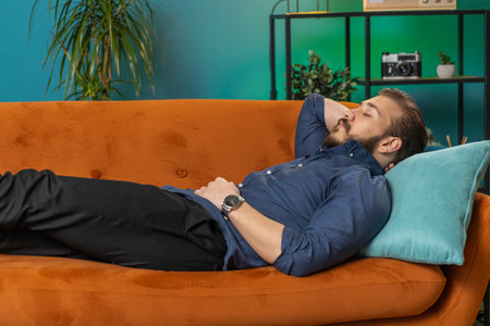 Tired lebanese man lying down on comfortable couch taking a rest, daytime nap. Portrait of carefree arabian guy holding hands behind head relaxing, falling asleep at home apartment living room on sofaの写真素材