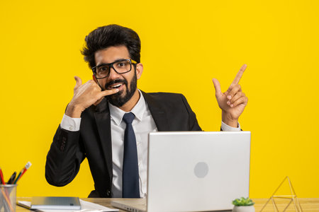 Call me, here is contact number. Indian businessman looking at camera doing phone gesture asking to make telephone conversation at office workplace desk. Hindu man in formal suit working on laptopの写真素材