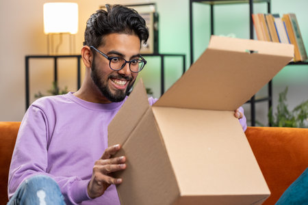 Happy indian man unpacking delivery parcel sitting at home apartment. Smiling satisfied hindu guy shopper, online shop customer opening cardboard box receiving purchase gift by fast postal shippingの写真素材