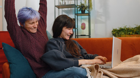 Two lesbian women family couple friends with laptop scream in delight clapping hands in triumph gesture celebrate success win money in lottery at home living room. Excited girls get online good newsの写真素材