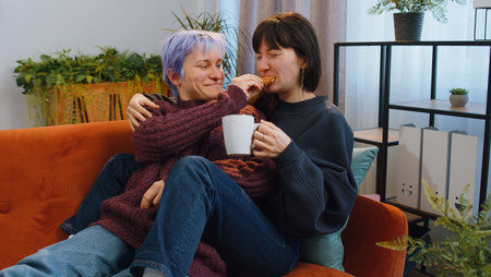 Two lesbian women family couple, girl friends drinking coffee or tea, eating croissants and talking at home morning living room. LGBT same-sex people. Enjoying happy breakfast together lying on sofaの写真素材