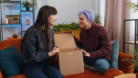 Happy two young lesbian women family couple unpacking delivery parcel at home living room. Smiling satisfied girls friends shoppers, online shop customers opening cardboard box receiving purchase giftの写真素材