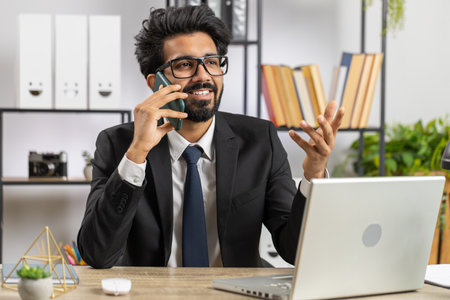 Bearded indian businessman working at office, having mobile phone talk at office desk with laptop....
