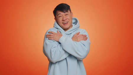 Asian handsome man spread hands and give hug to you. Pleasant expression, love feelings. Adult guy isolated alone on orange studio background. People lifestylesの写真素材