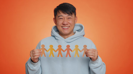 Man showing multiracial different skin color human shape cut out of paper, concept of teamwork, partnership, community, unity, people support love against racism. Asian guy on orange studio backgroundの写真素材