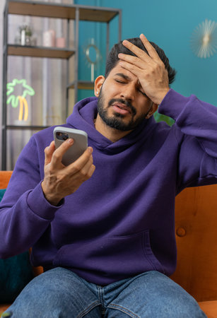 Sad displeased indian man use smartphone typing browsing, loses becoming surprised sudden lottery results, bad news, fortune loss, fail, deadline, virus. Young hindu guy at home living room on couchの写真素材
