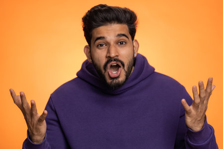 Oh my God, Wow. Excited amazed winner man raising hands in surprise looking at camera with big eyes, shocked by sudden victory. Sincere handsome indian guy isolated alone on orange studio backgroundの写真素材