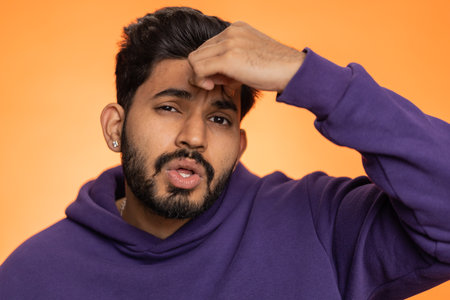 Handsome indian bearded man in hoodie rubbing temples to cure headache problem, suffering from tension, migaine, stress, grimacing in pain, high blood pressure isolated alone on orange wall backgroundの写真素材