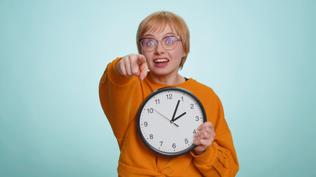 It is your time. Young woman in glasses showing time on wall office clock, ok, thumb up, approve,...