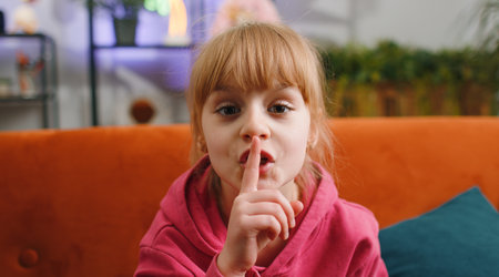 Shh be quiet please. Young blonde child kid girl presses index finger to lips makes silence hush gesture sign do not tell gossip privacy secret. Female teen toddler at home in living room sits on sofaの写真素材