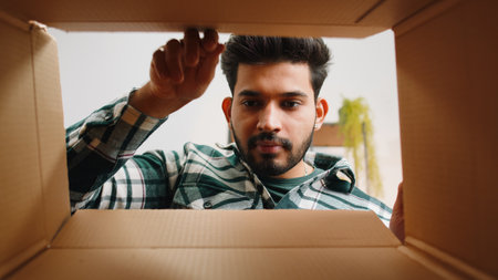 Inside view from box. Indian man unpacking delivery parcel at home. Smiling satisfied hindu guy...