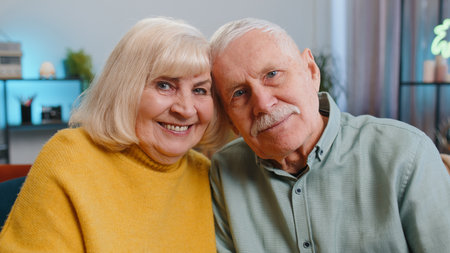 Happy old senior elderly family couple hugging, laughing, smiling looking at camera, healthy retired two grandparents husband wife. Grandmother grandfather embracing sitting on sofa at home, portraitの写真素材