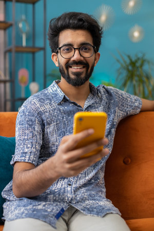 Indian man sitting on sofa uses mobile phone smiles at home living room apartment. Hindu young guy...