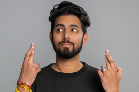 Indian young man crossed fingers looking at camera asking for good lucky news, wishing good exam results, dreaming about win victory in lottery jackpot. Hindu guy isolated on gray studio backgroundの写真素材