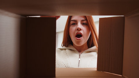 Inside view from box. Happy redhead woman unpacking delivery parcel at home. Smiling satisfied girl shopper online shop customer opening cardboard receiving purchase gift by fast postal shipping orderの写真素材