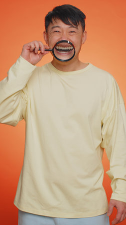 Asian man holding magnifier glass on healthy white teeth, looking at camera with happy expression, showing funny silly face smiling mouth. Chinese guy isolated on orange background. Verticalの写真素材