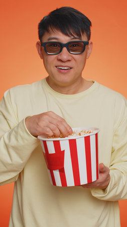 Excited asian man in 3D glasses eating popcorn, watching interesting TV serial, sport game, film,...
