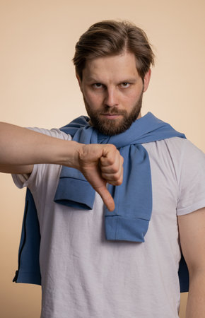 Dislike. Upset unhappy caucasian man in t-shirt showing thumbs down sign gesture, expressing discontent, disapproval, dissatisfied, dislike. Handsome young guy. Indoor studio shot on beige backgroundの写真素材