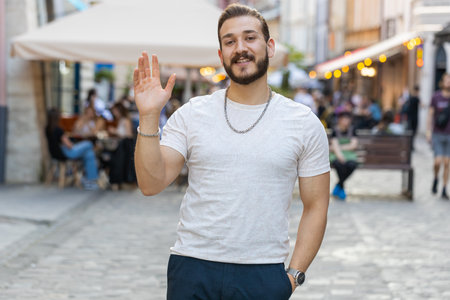 Portrait of bearded young man smiling friendly at camera, waving hands gesturing hello, hi, greeting or goodbye, welcoming with hospitable expression outdoors. Guy walking in urban city town streetの写真素材