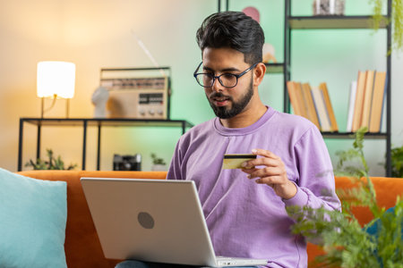Young indian man using credit bank card and laptop computer while transferring money, purchases online shopping, order food delivery. Portrait of hindu guy at home apartment living room on couchの写真素材