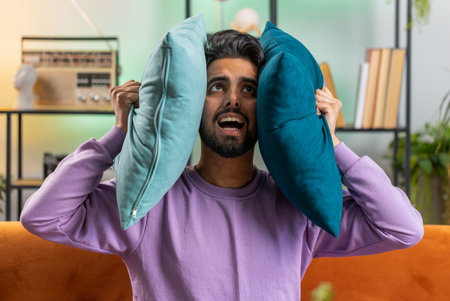 Repair work at neighbours. Irritated indian man relaxing on couch cover ears with pillows annoyed by noisy neighbors suffer from headache wish silence. Thin walls at home flat without sound insulationの写真素材