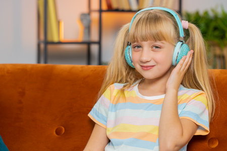 Happy overjoyed toddler girl kid in wireless headphones relaxing sitting on couch at home room choosing listening favorite energetic disco dancing music. Child weekends leisure activities. Lifestylesの写真素材